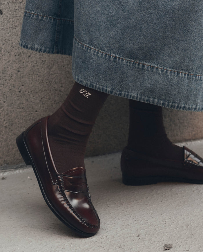 A person wearing THE GREAT. F26 D1 The Monogrammed Trouser Sock in Chocolate, featuring embroidered initials "T.G.", along with dark brown loafers and a denim skirt, standing on a gray surface.