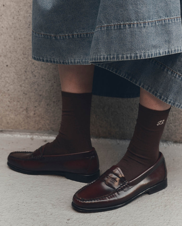 A close-up of a person's lower legs wearing THE GREAT. F26 D1 The Monogrammed Trouser Sock in Chocolate, featuring embroidered "T.G." initials, styled with dark brown leather loafers and a denim skirt, standing on a light-colored surface.