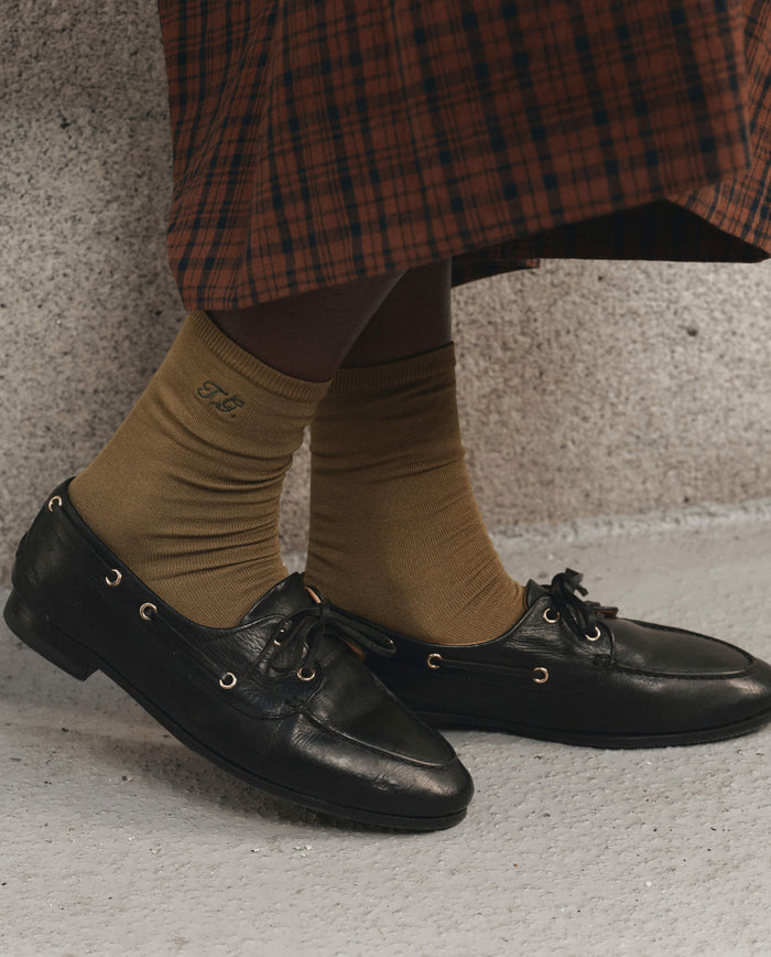 A person wearing THE GREAT. F26 D1 The Monogrammed Trouser Sock in Golden Ochre and black leather loafers stands on a light-colored floor, dressed in a brown and black checked skirt.