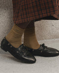 A person wearing THE GREAT. F26 D1 The Monogrammed Trouser Sock in Golden Ochre and black leather loafers stands on a light-colored floor, dressed in a brown and black checked skirt.