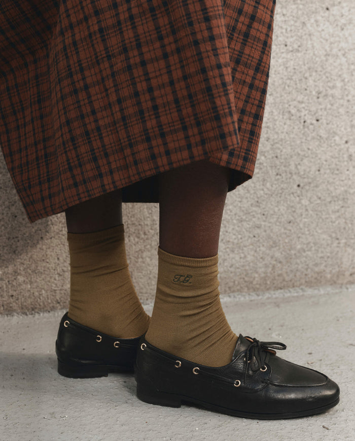 A person stands on a concrete surface wearing THE GREAT. F26 D1's The Monogrammed Trouser Sock in Golden Ochre, personalized with the initials "TC," paired with black loafers and a brown plaid skirt.