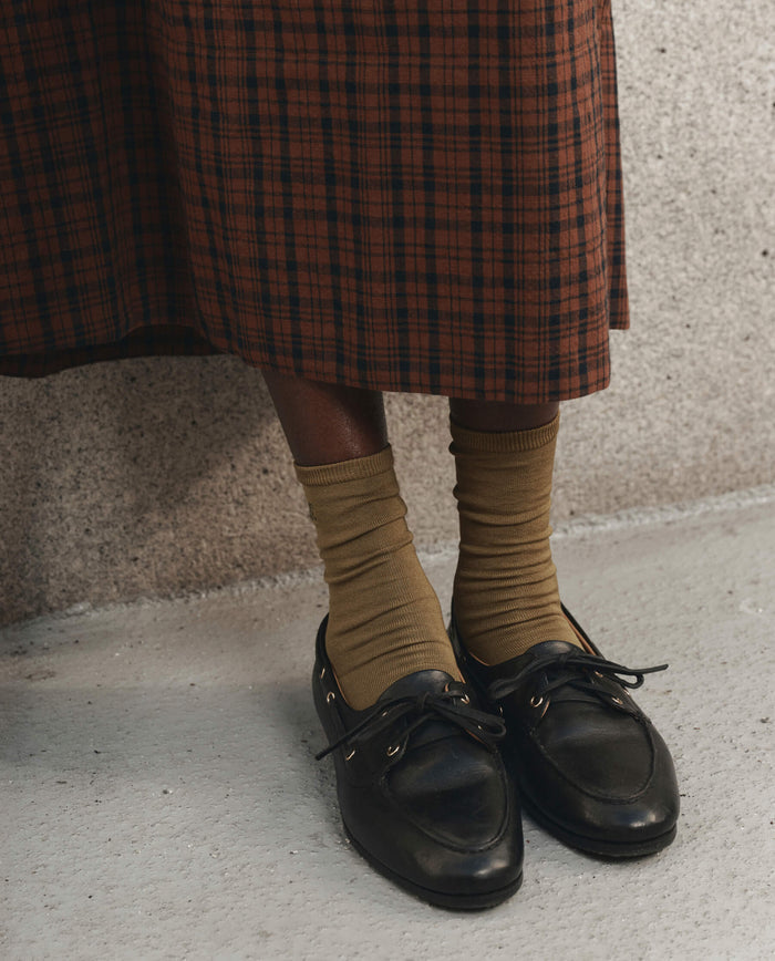 A person wearing a brown plaid skirt, THE GREAT. F26 D1 The Monogrammed Trouser Sock in Golden Ochre, and black loafers stands on a concrete floor near a textured wall.