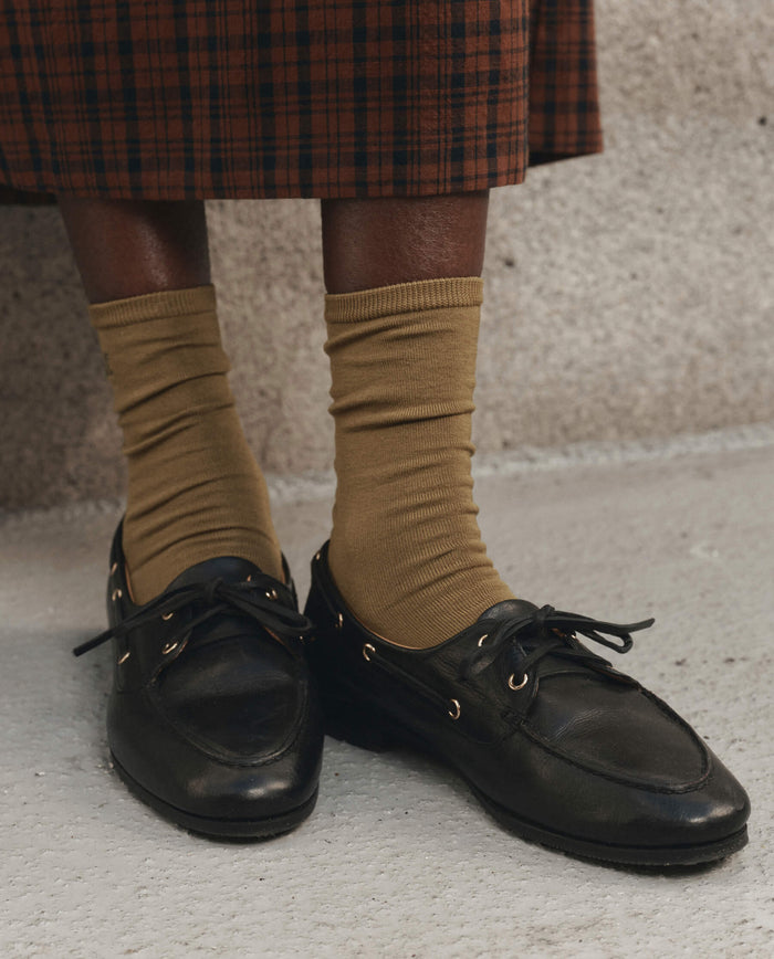 A person wearing THE GREAT. F26 D1 The Monogrammed Trouser Sock in Golden Ochre and black leather loafers stands on a concrete surface, with a brown plaid skirt visible in the upper part of the image.