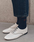 A person stands on a gray concrete surface wearing cropped dark blue jeans, white lace-up shoes, and THE GREAT. F26 D1's The Monogrammed Trouser Sock in Navy, featuring red embroidery.