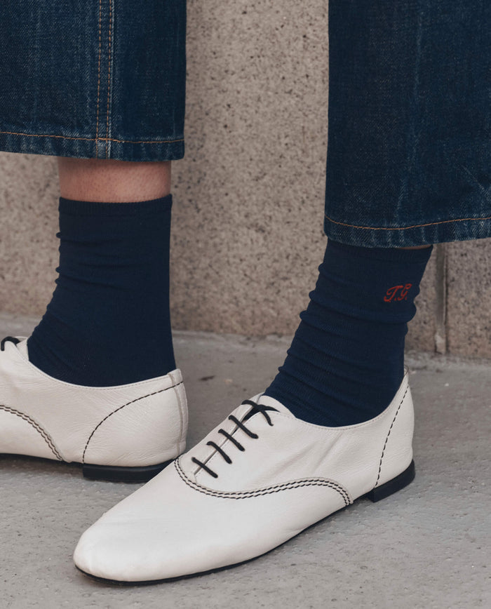 A person wearing blue jeans, THE GREAT. F26 D1 The Monogrammed Trouser Sock in navy with orange initials, and white lace-up leather shoes with black stitching.