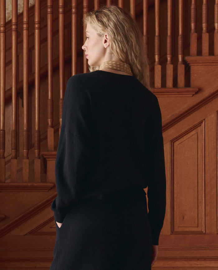 A person with wavy blonde hair, wearing THE GREAT. SP26 D1 The Oversized Pullover in black, stands indoors facing away from the camera by wooden stairs and accessorized with a tan scarf.