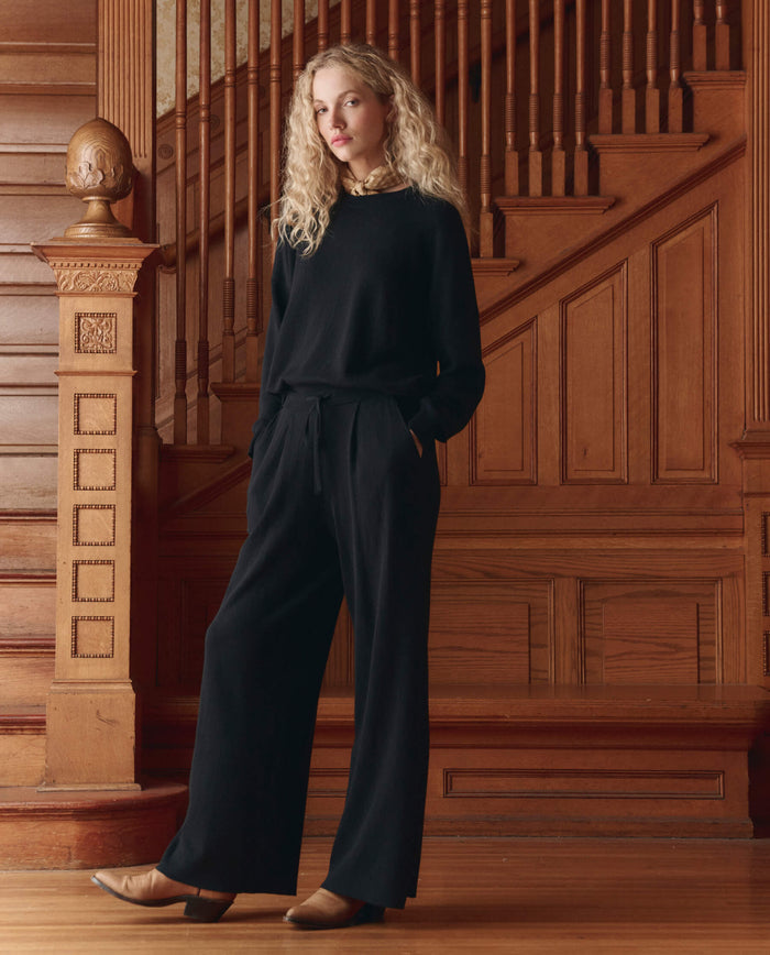A person with long curly blonde hair stands by a wooden staircase, wearing THE GREAT. SP26 D1 The Oversized Pullover in black, paired with black trousers and tan shoes.