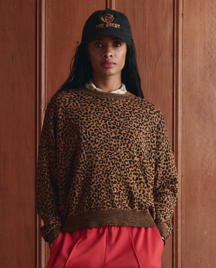 A person in a black cap, red pants, and THE GREAT. SP26 D1 The Oversized Pullover -- Leopard stands before wooden paneling, looking at the camera.