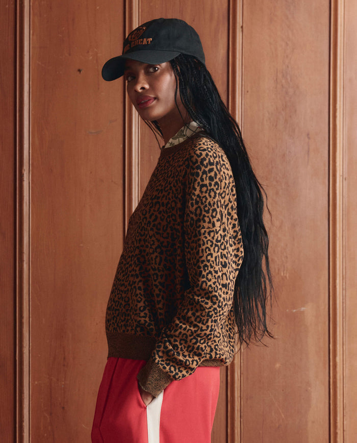 A person wearing THE GREAT. SP26 D1 The Oversized Pullover in leopard and a black cap with red pants stands sideways before a wooden panel wall.