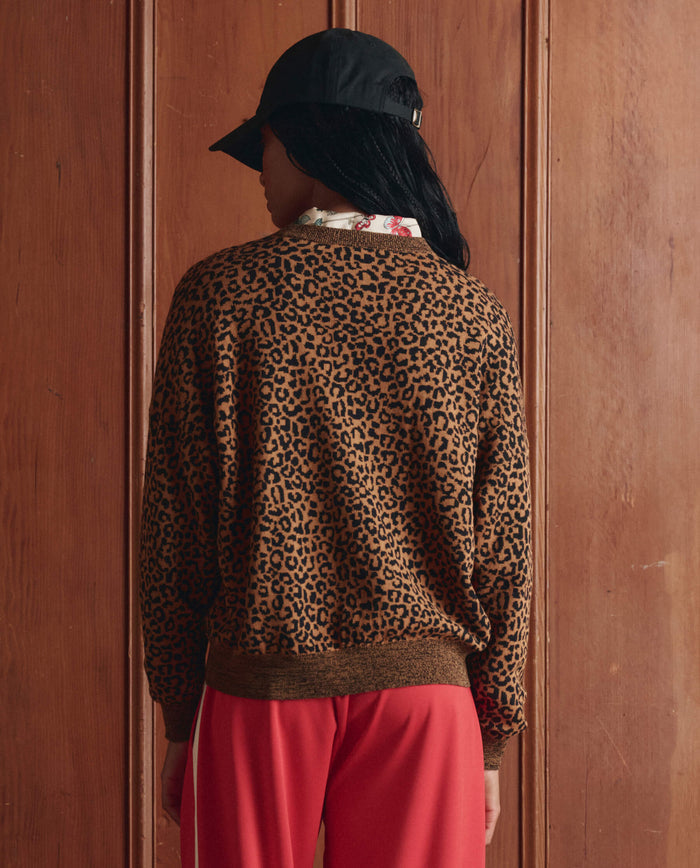 A person stands with their back to the camera, facing a wooden-paneled wall, wearing a black cap, red pants, and THE GREAT. SP26 D1 The Oversized Pullover in leopard print.