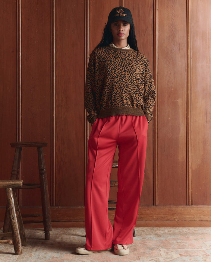 Indoors, a person wears THE GREAT. SP26 D1 The Oversized Pullover in Leopard, red wide-leg pants, a black cap, and beige shoes, hands in pockets. Wooden stools are nearby.