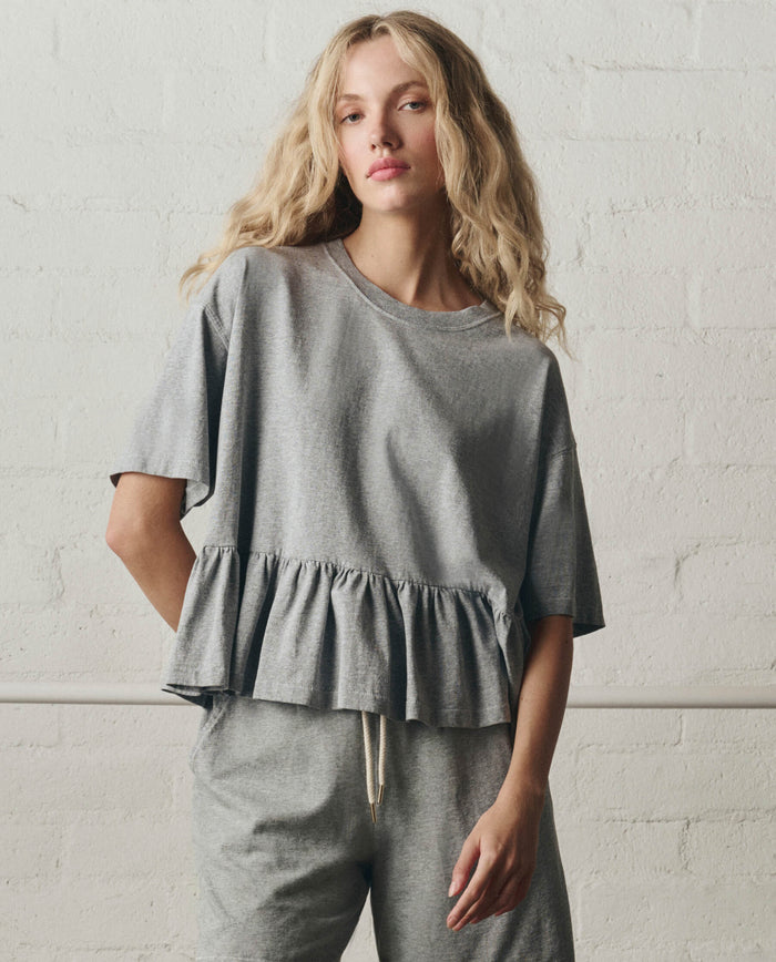 A woman stands against a white brick wall wearing THE GREAT.’s The Oversized Ruffle Peplum Tee in Heather Grey with matching pants.