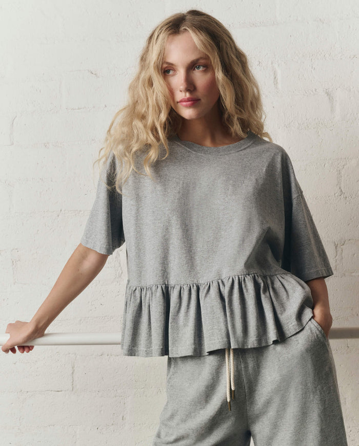 Woman with long wavy blonde hair wears THE GREAT. SP26 DANCER BODIES' The Oversized Ruffle Peplum Tee in Heather Grey and matching pants, standing against a white brick wall.