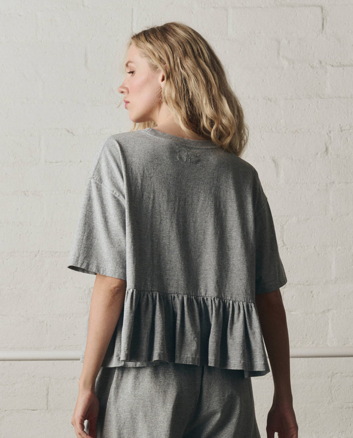 A woman stands with her back to the camera in front of a white brick wall, wearing THE GREAT. SP26 DANCER BODIES' The Oversized Ruffle Peplum Tee in Heather Grey with matching grey bottoms.