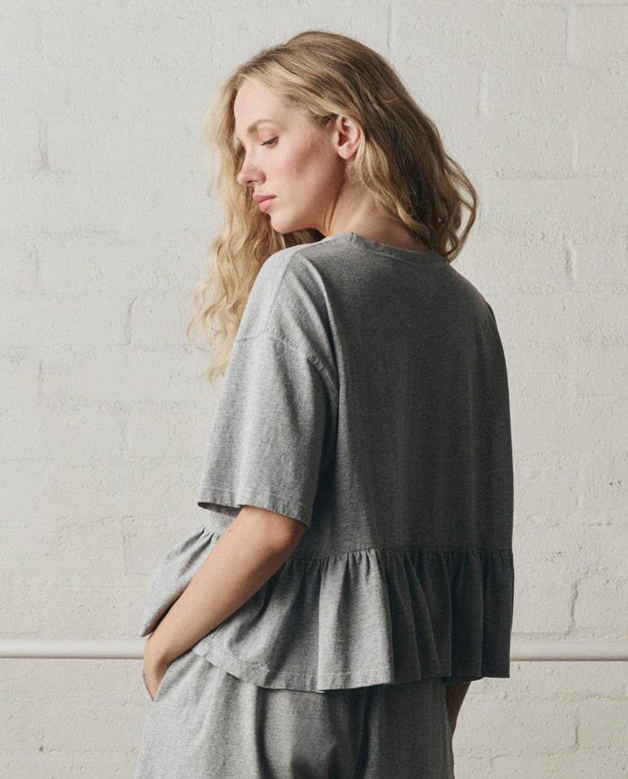 A woman with long blonde hair stands sideways against a white brick wall, wearing THE GREAT.'s Oversized Ruffle Peplum Tee in Heather Grey from SP26 DANCER BODIES.