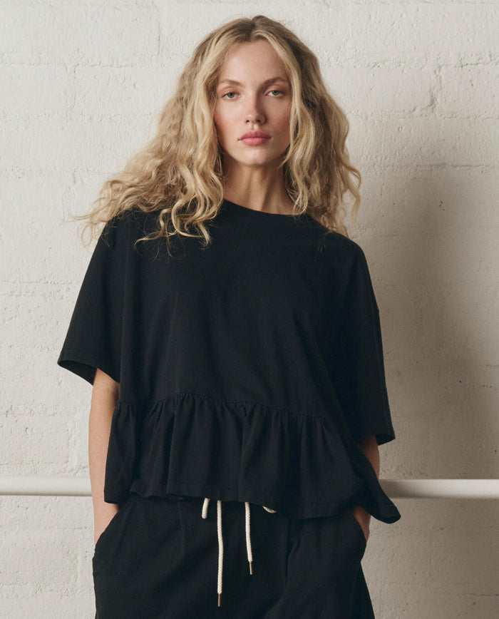 A woman with long wavy blonde hair wears THE GREAT. SP26 DANCER BODIES The Oversized Ruffle Peplum Tee in True Black and black drawstring pants, standing against a white textured wall.