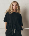 A woman with long wavy blonde hair wears THE GREAT. SP26 DANCER BODIES The Oversized Ruffle Peplum Tee in True Black and black drawstring pants, standing against a white textured wall.