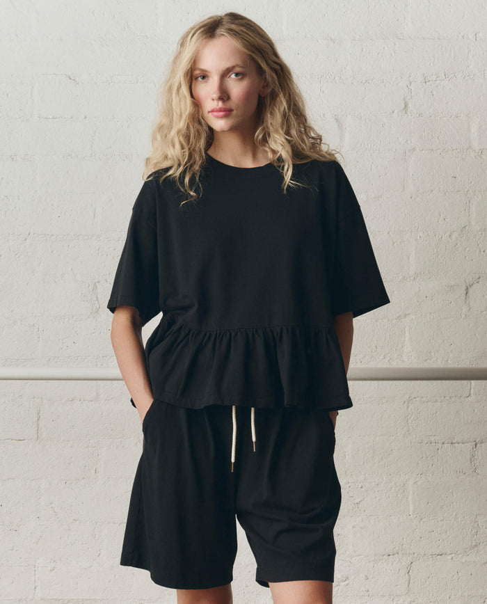 A person stands against a white brick wall wearing THE GREAT. SP26 DANCER BODIES The Oversized Ruffle Peplum Tee in True Black with matching black shorts, hands in pockets.