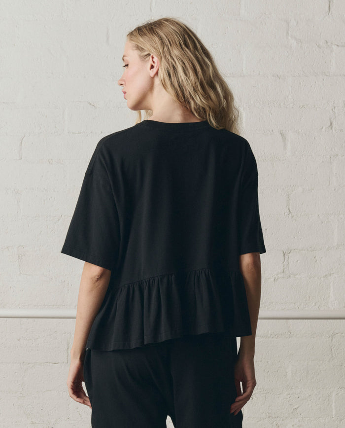 A person with long blonde hair stands against a white brick wall, wearing THE GREAT. SP26 DANCER BODIES’ The Oversized Ruffle Peplum Tee in True Black.