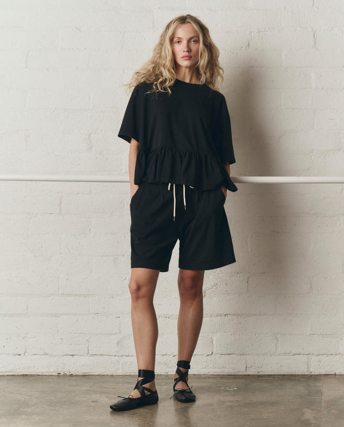 A person stands indoors against a white brick wall, wearing THE GREAT. SP26 DANCER BODIES' The Oversized Ruffle Peplum Tee in True Black, styled with drawstring black shorts and black ankle-strap ballet flats.