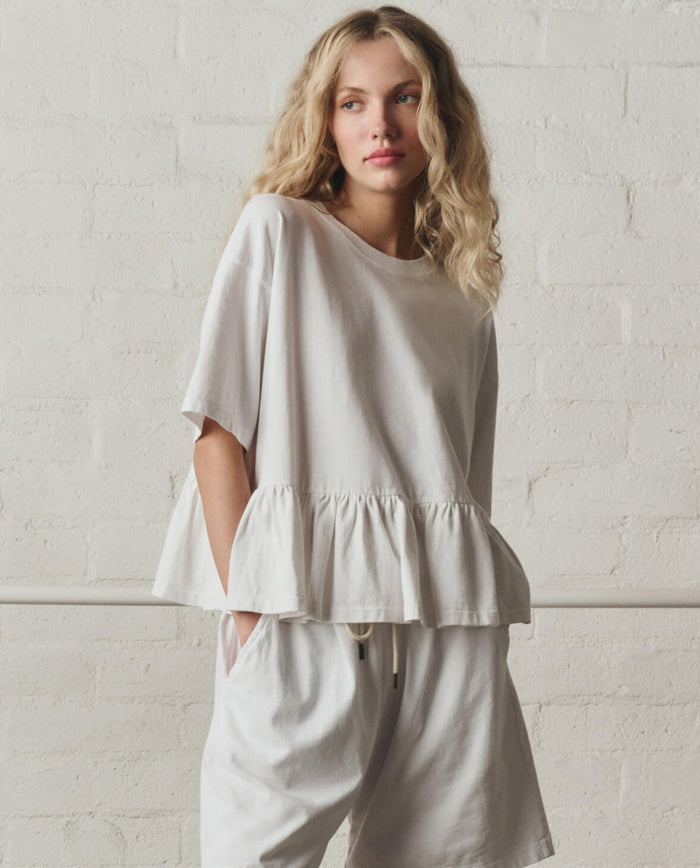 A woman with wavy blonde hair wears THE GREAT.'s Oversized Ruffle Peplum Tee in True White and matching shorts, posing against a white brick wall.