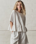 A woman with wavy blonde hair wears THE GREAT.'s Oversized Ruffle Peplum Tee in True White and matching shorts, posing against a white brick wall.