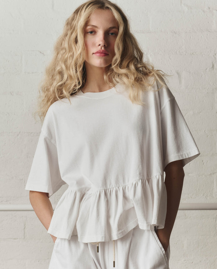 A person stands against a white brick wall wearing THE GREAT.'s The Oversized Ruffle Peplum Tee in True White, styled with matching white pants.