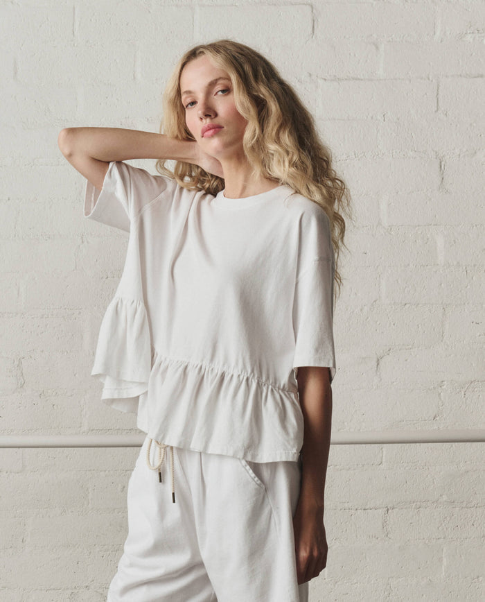 A woman stands against a white brick wall in THE GREAT. SP26 DANCER BODIES The Oversized Ruffle Peplum Tee in True White and matching pants, her right arm raised to touch her hair.