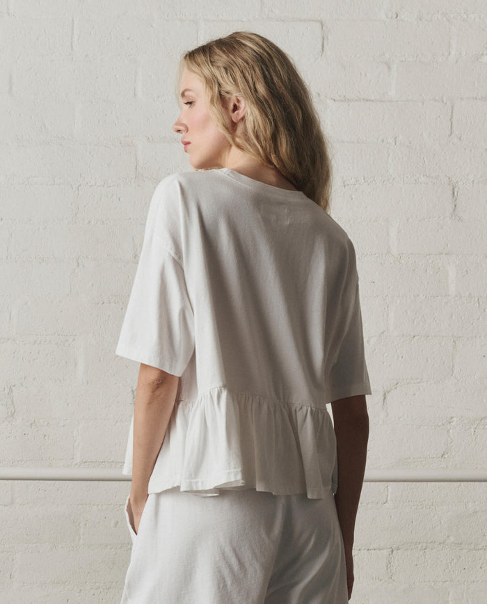 A person with long blonde hair faces away, wearing THE GREAT.’s The Oversized Ruffle Peplum Tee in True White with matching pants, standing against a white brick wall.