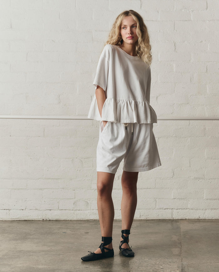 A woman stands indoors against a white brick wall, wearing THE GREAT.'s The Oversized Ruffle Peplum Tee in True White from the SP26 DANCER BODIES line, paired with matching shorts and black ankle-tie flats.
