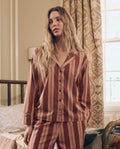 A person with long wavy hair stands in a bedroom, wearing The Pajama Shirt -- Ganache Stripe by THE GREAT. F26 SLEEP, a striped loungewear set in shades of brown and pink. A bed and patterned curtains are visible in the background.