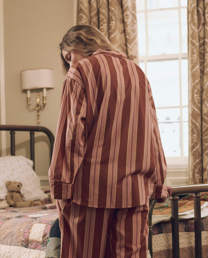 Wearing THE GREAT. F26 SLEEP’s The Pajama Shirt in Ganache Stripe, a person sits on the edge of a bed in a cozy, warmly lit bedroom with patterned curtains and a teddy bear resting on the bed.