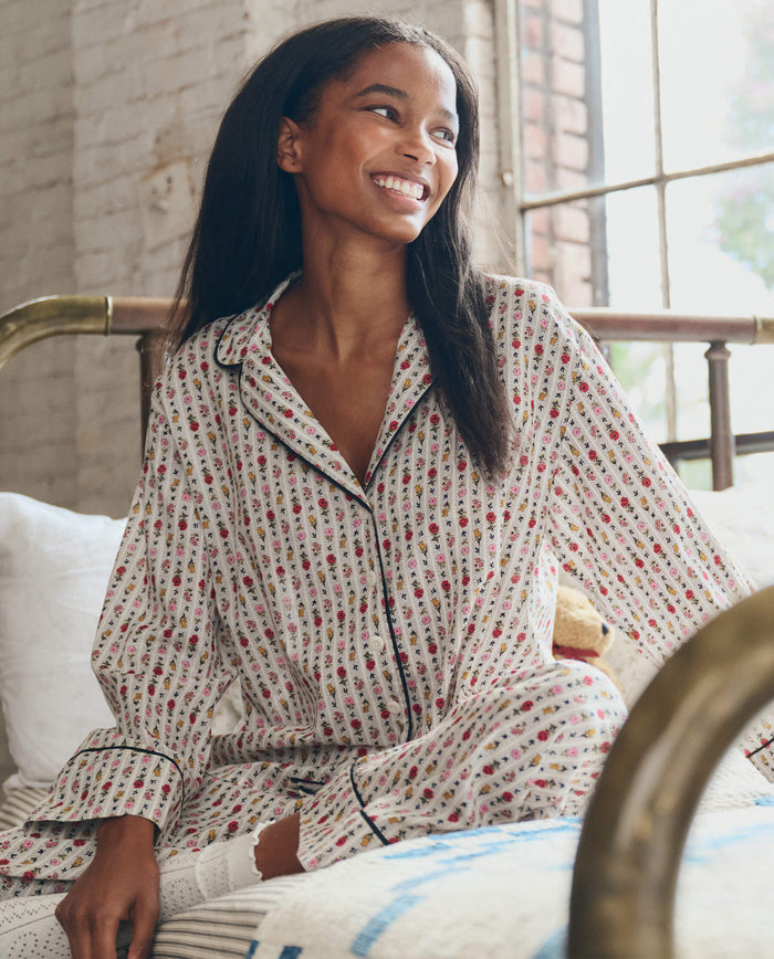 A woman in The Pajama Shirt -- Ribbon Floral by THE GREAT. PS26 SLEEP relaxes on a bed, enjoying the comfort of this classic cotton sleepwear.