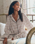 A woman in The Pajama Shirt -- Ribbon Floral by THE GREAT. PS26 SLEEP relaxes on a bed, enjoying the comfort of this classic cotton sleepwear.