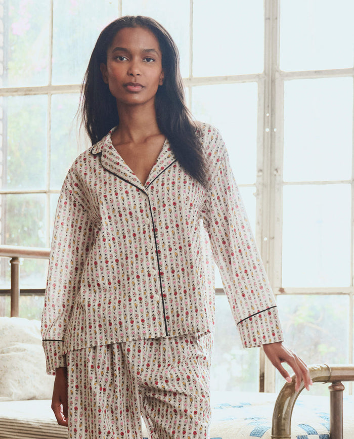 A woman stands indoors in THE GREAT. PS26 SLEEP The Pajama Shirt -- Ribbon Floral, a matching patterned cotton pajama set with a collared button-up shirt and long pants, illuminated by natural light from large windows.