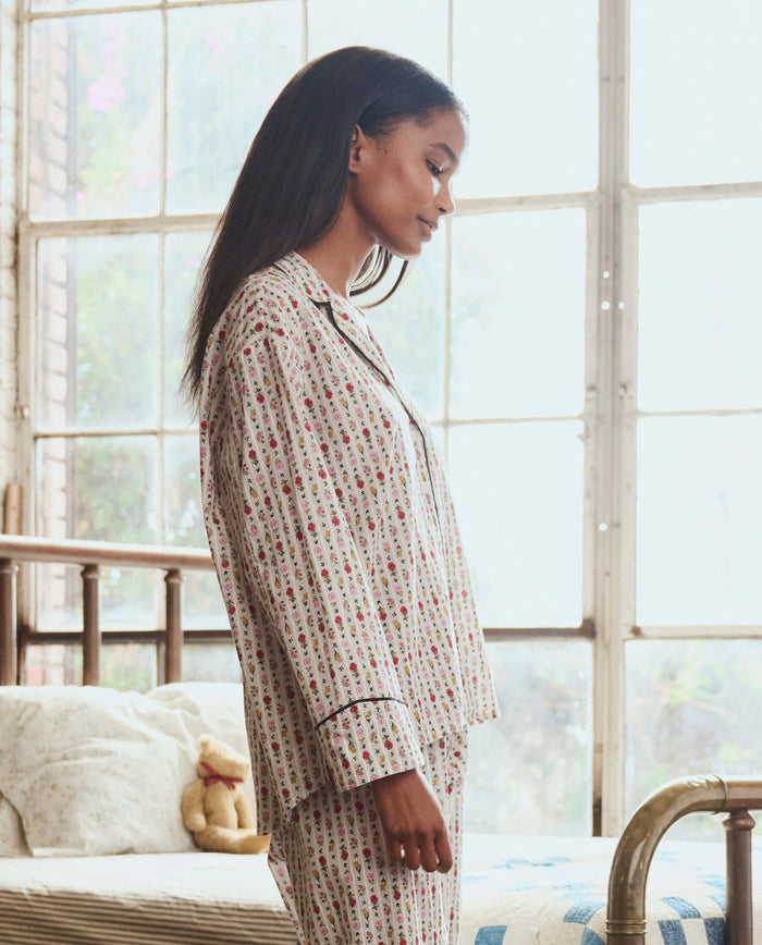 A woman wearing THE GREAT. PS26 SLEEP "The Pajama Shirt" in Ribbon Floral stands sideways in a sunlit bedroom with large windows, a bed, and a stuffed animal in the background.