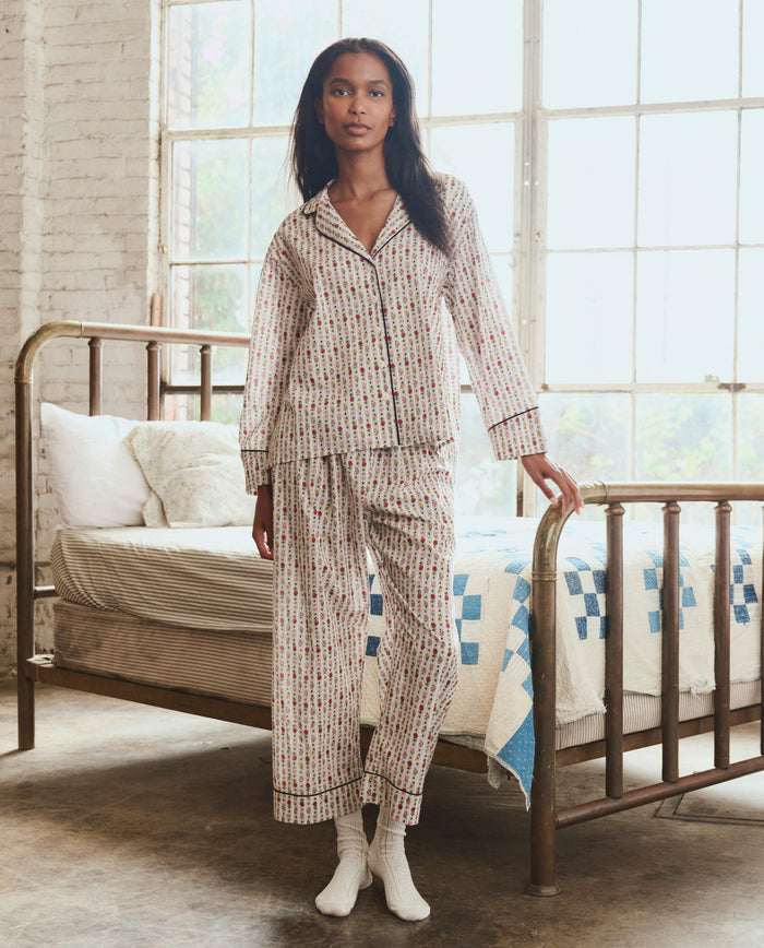 A woman stands in a sunlit bedroom wearing THE GREAT. PS26 SLEEP The Easy Sleep Pant in Ribbon Floral, with one hand on a brass bed frame and a quilt visible behind her.