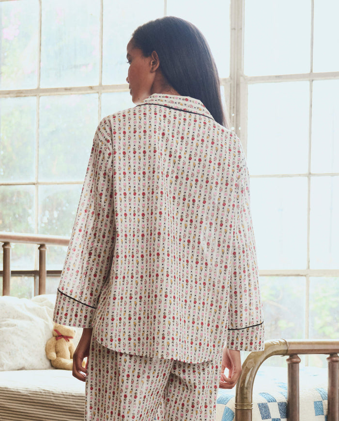 A woman stands indoors facing away, wearing THE GREAT. PS26 SLEEP The Pajama Shirt in Ribbon Floral, with large windows behind her and a bed with a stuffed animal visible in the background.
