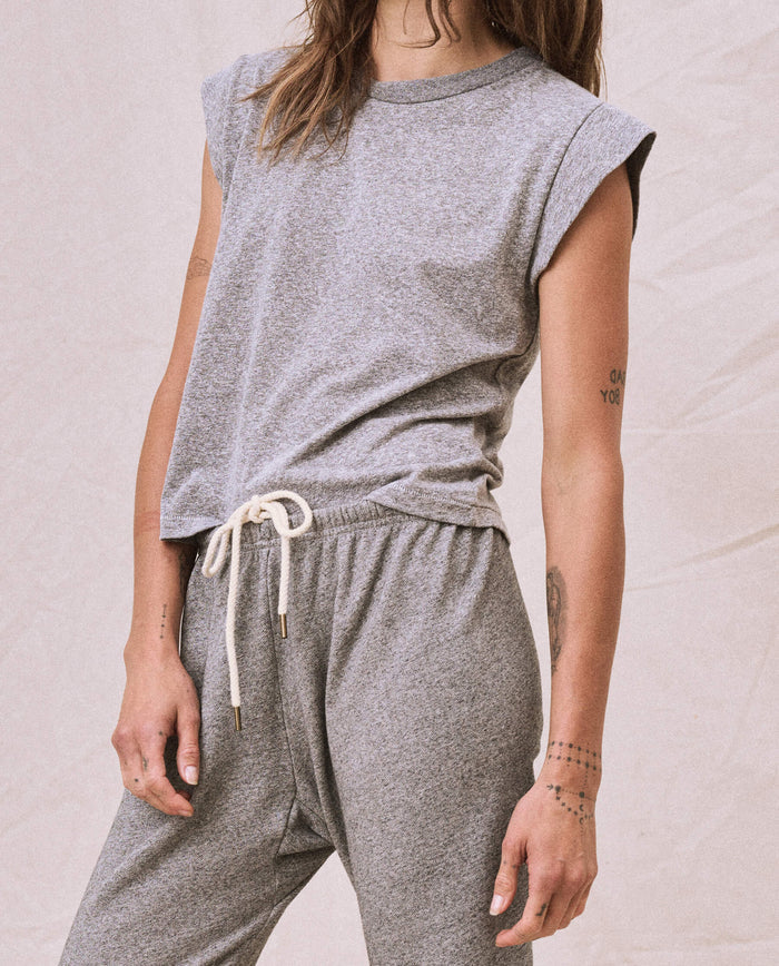 A person wearing a heather grey Peak Shoulder Tee from THE GREAT. FALL 22 KNITS, paired with matching high-waisted sweatpants featuring white drawstrings, stands against a light-colored background. Their arms are adorned with multiple tattoos.