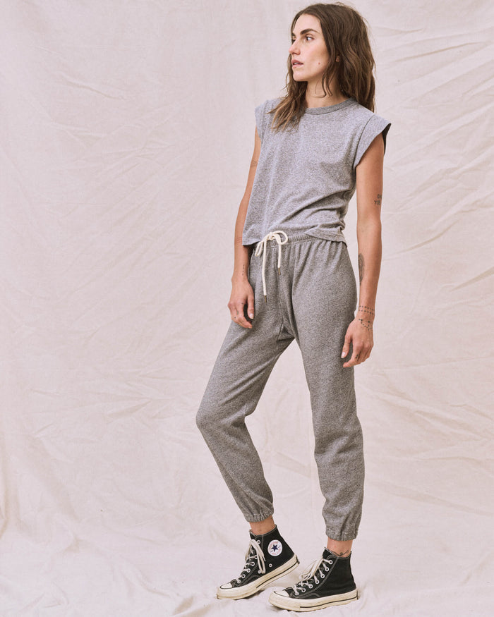 A woman stands against a plain fabric backdrop, wearing The Peak Shoulder Tee in heather grey by THE GREAT. FALL 22 KNITS and matching drawstring joggers, complemented by black high-top sneakers. She looks to her left with a relaxed posture.