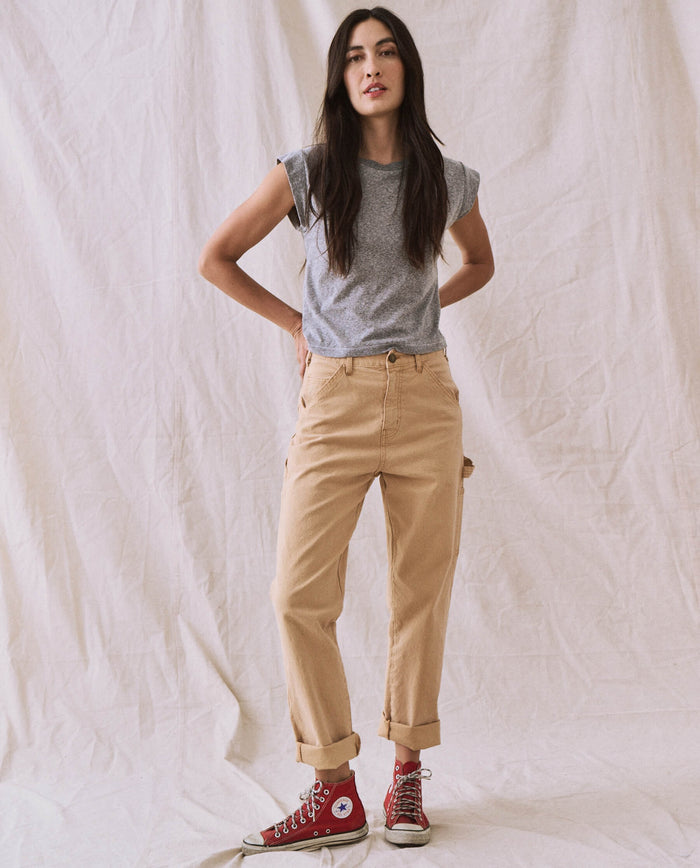 A person stands against a white fabric backdrop, wearing The Peak Shoulder Tee in Heather Grey by THE GREAT. FALL 22 KNITS, beige rolled-up pants, and red high-top sneakers. Their hands are on their hips.