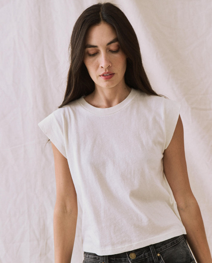A woman with long, dark hair stands against a light background wearing a handcrafted, locally made Peak Shoulder Tee in Washed White from THE GREAT. FALL 22 KNITS and dark high-waisted denim. She is looking downward with a neutral expression.