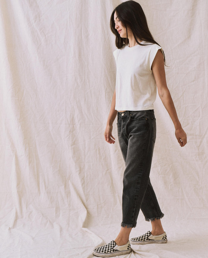 A person with long hair, wearing a handcrafted locally-made Peak Shoulder Tee in washed white from THE GREAT. FALL 22 KNITS, high-waisted denim, and checkered slip-on shoes walks against a neutral fabric backdrop.