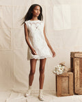 A woman in THE GREAT. PS26's The Picnic Shift Dress -- Cream stands by wooden crates and white flowers against a neutral backdrop; her look is elegant yet casual with crochet lace details and white sneakers.