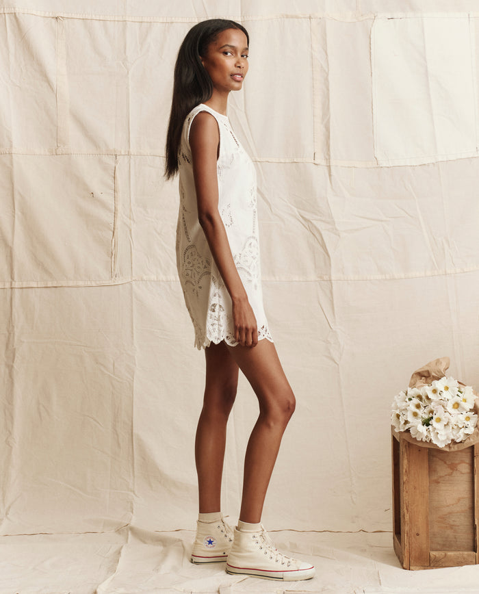 A woman in THE GREAT. PS26 The Picnic Shift Dress in cream and white high-top sneakers stands sideways before a beige fabric backdrop, next to a wooden crate adorned with white flowers.