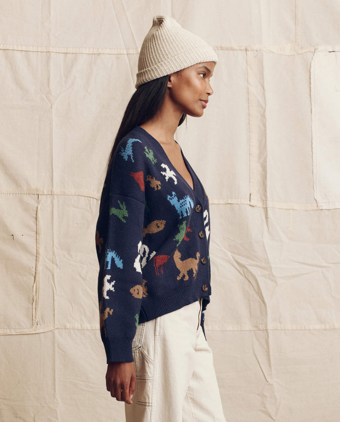 A woman in a cream knit beanie, white pants, and THE GREAT. PS26 The Pictograph Slouch Cardigan in Navy stands in profile before a beige fabric backdrop.