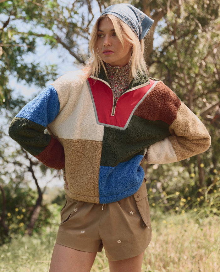 A person stands outdoors in THE GREAT. SP26 TGO's Plush Color Block Sierra Pullover in Canyon Multi, paired with tan floral shorts and a blue headscarf, surrounded by trees and grass.
