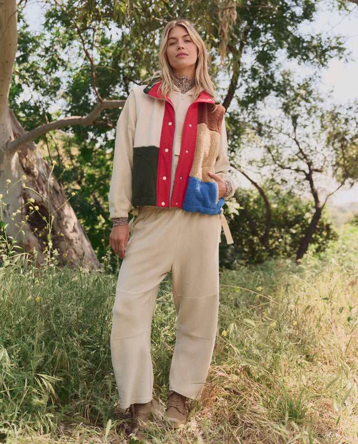 A person stands outdoors amid trees and tall grass, wearing a beige outfit and THE GREAT.'s The Plush Color Block Sierra Vest in Canyon Multi.