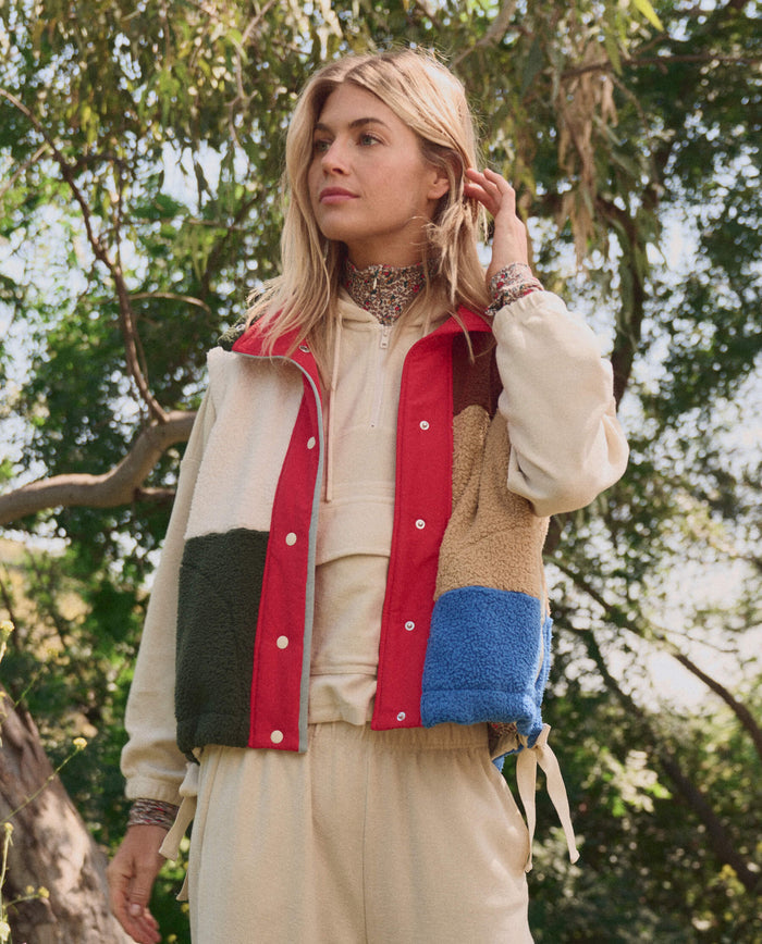 A woman stands outdoors among trees, wearing THE GREAT. SP26 TGO The Plush Color Block Sierra Vest in Canyon Multi over a beige tracksuit.
