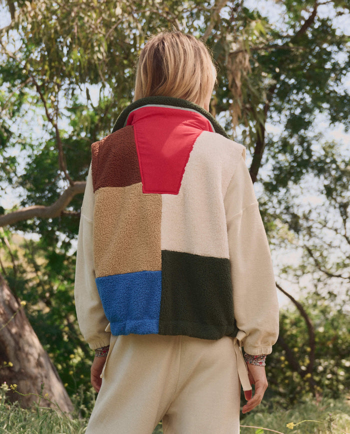 A person stands outdoors among greenery, wearing a cream outfit and THE GREAT. SP26 TGO The Plush Color Block Sierra Vest in Canyon Multi—featuring red, brown, blue, green, and white color blocks.